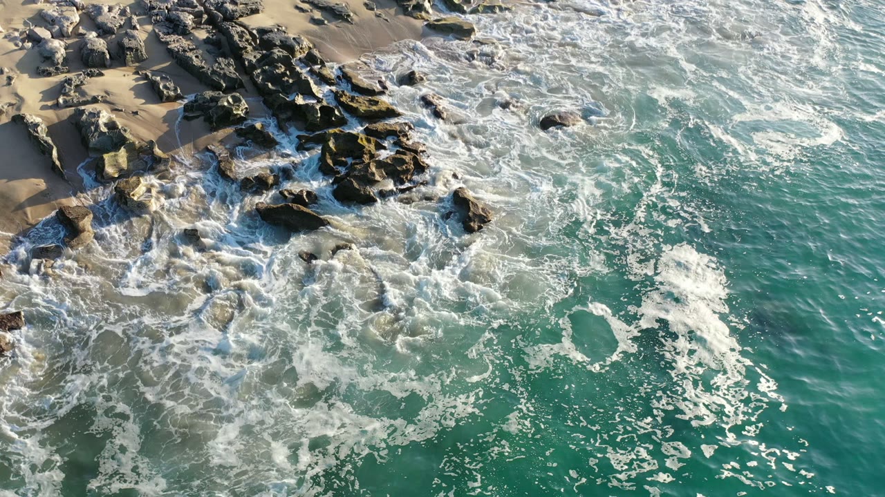 Aerial frame from Los Cabos, Mexico showing green-blue ocean water breaking into white foam around dark shoreline rocks and pale sand, with no visible people.
