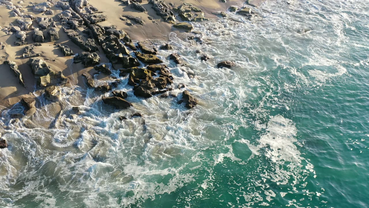 Aerial Los Cabos shoreline view with whitewater spreading over the rocks at left and clearer green ocean filling the lower right, with no visible people.
