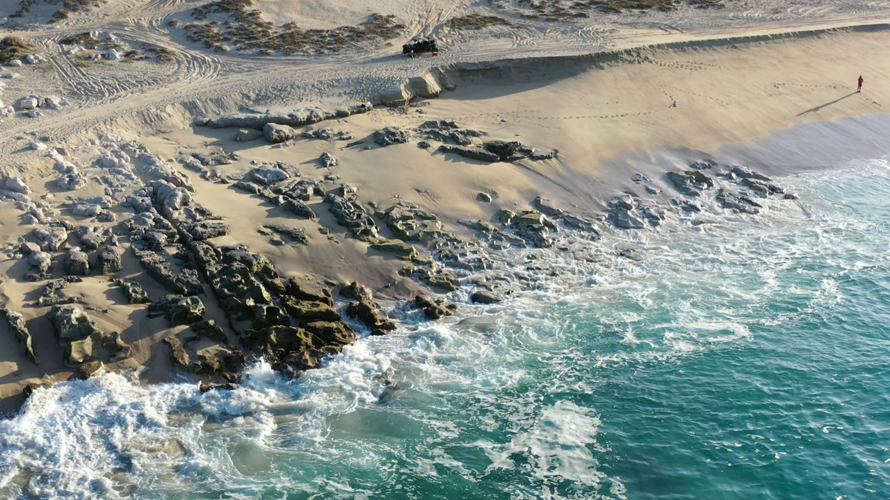 Aerial frame from Los Cabos, Mexico showing turquoise waves breaking around dark shoreline rocks, pale sand, dune tracks, low scrub, and no visible people.