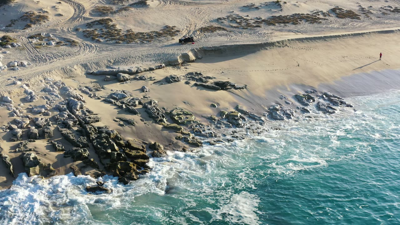 Aerial view in Los Cabos, Mexico with white foam spreading wider across turquoise water beside the rock cluster and empty sand, with no visible people.