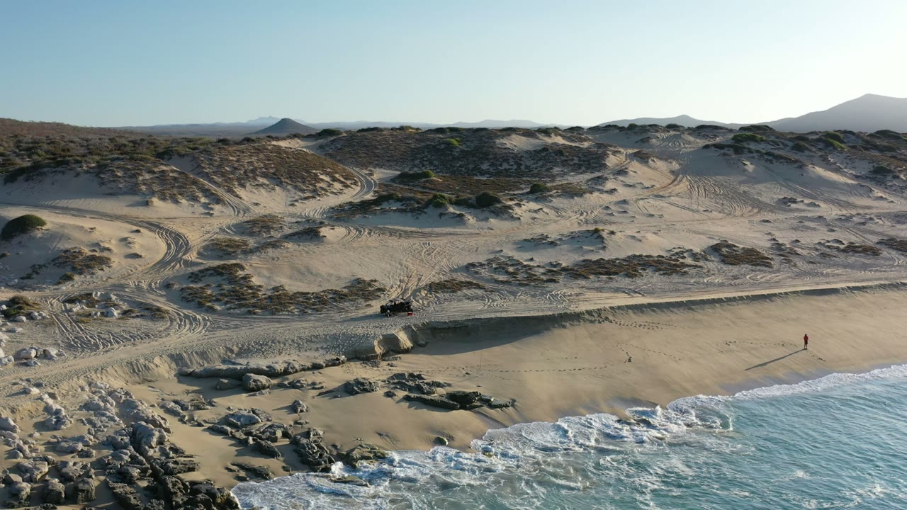 Wide aerial view of sandy coastal dunes, low scrub, rocks, and surf.