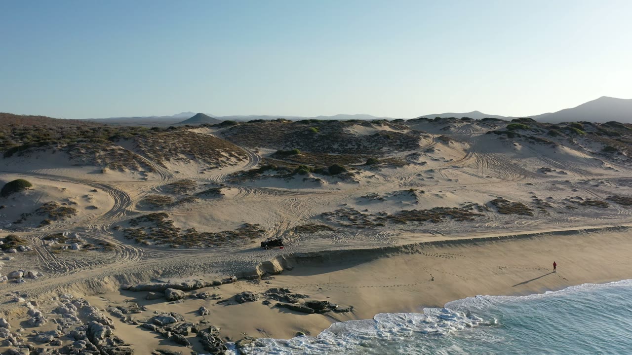 A Los Cabos aerial view shows curving tire tracks through sandy dunes, scattered scrub, rocky beach texture, and white waves reaching the lower frame, with no identifiable people visible.