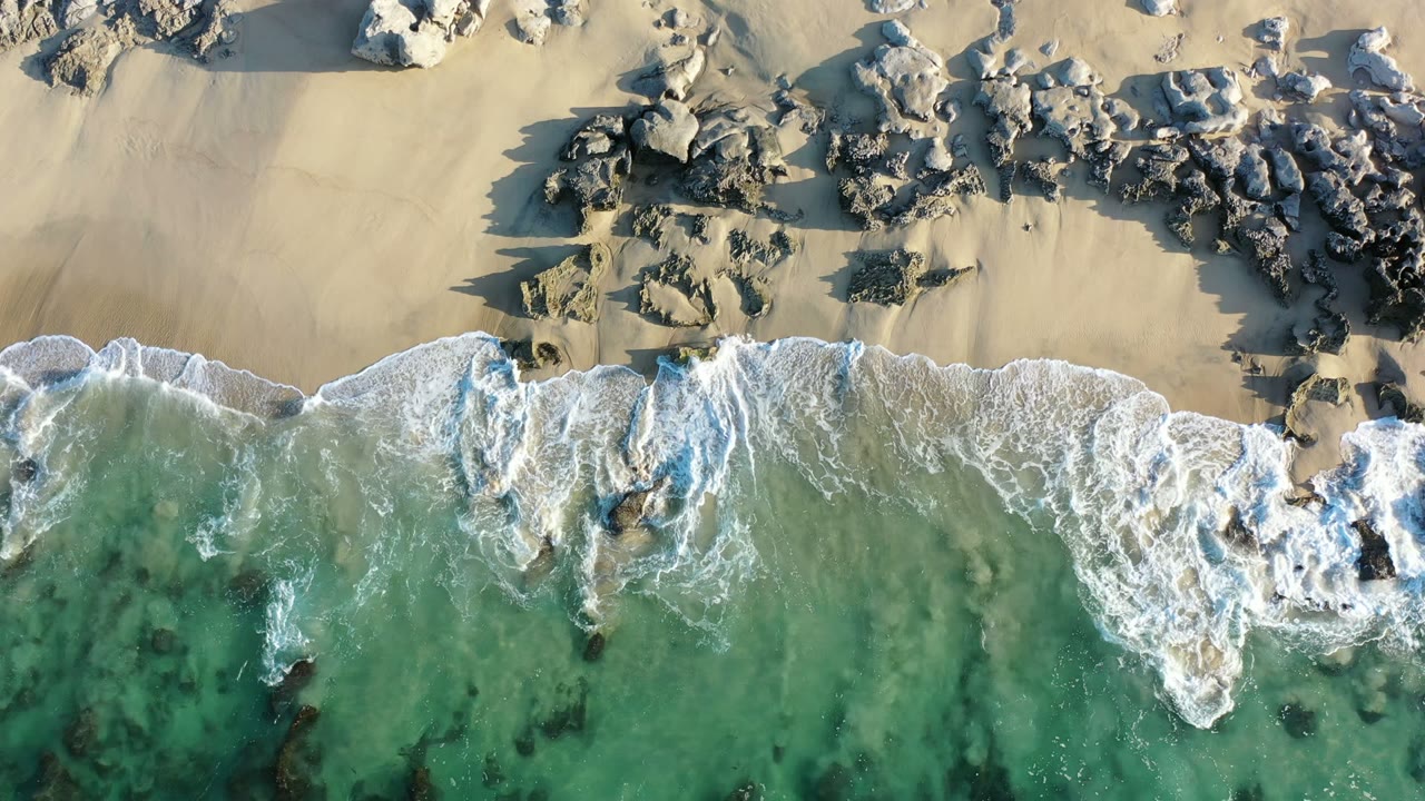 Aerial frame of white foam spreading around nearshore rocks as green water reaches the sandy Los Cabos beach, with no visible people.