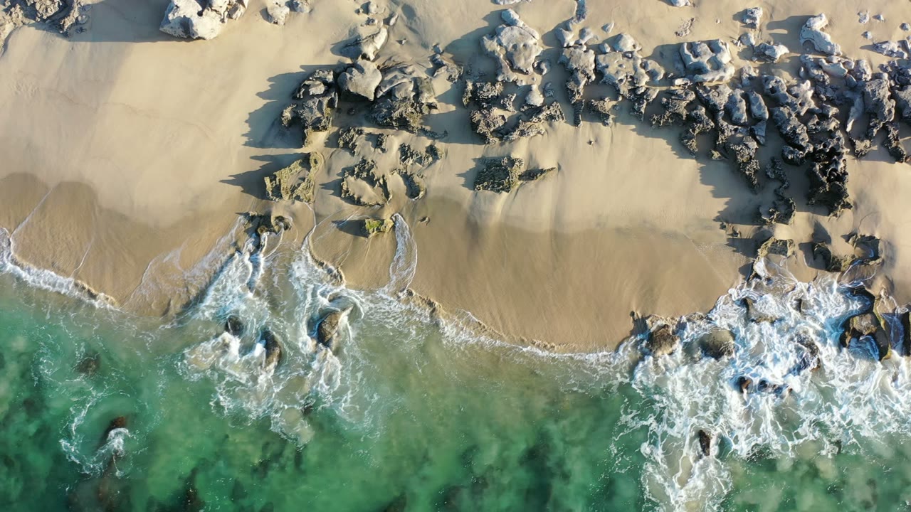 Overhead view of a Los Cabos beach where green-blue water pushes white foam around dark rocks on pale sand, with no visible people.
