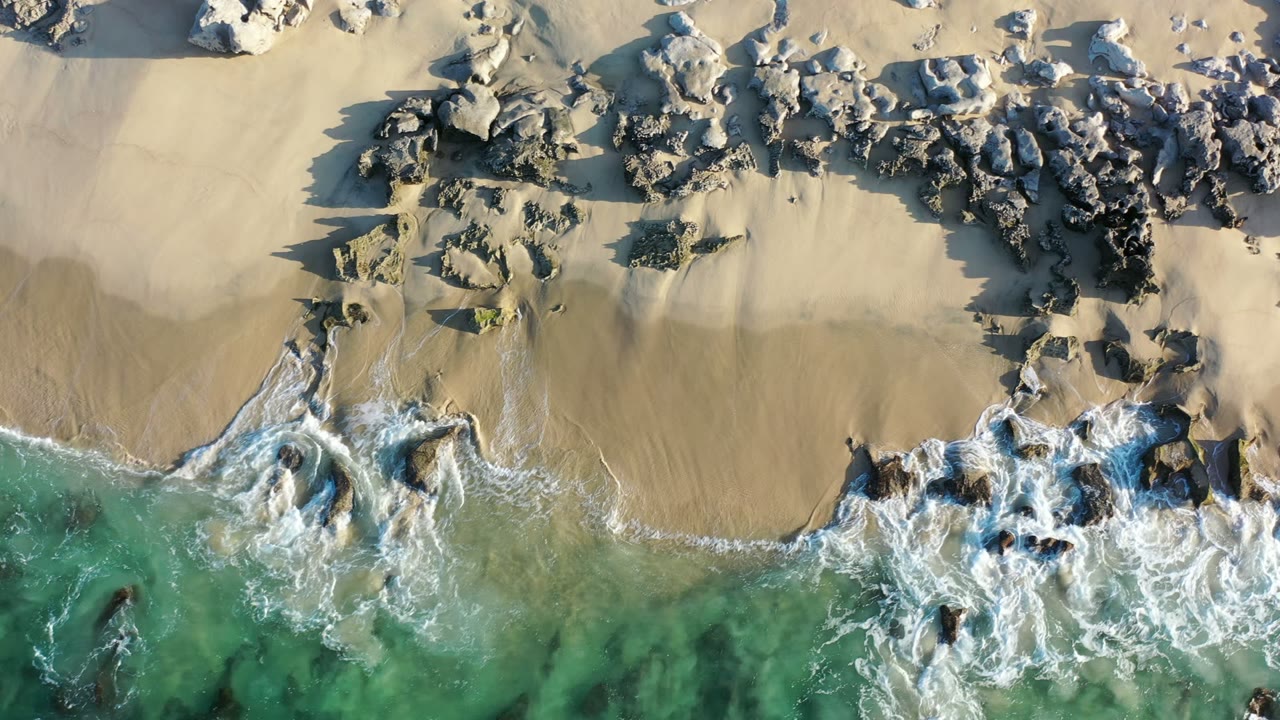 Top-down Los Cabos shoreline with a larger central sand area, clustered rocks above the wash line, and white foam tracing the lower beach, with no visible people.
