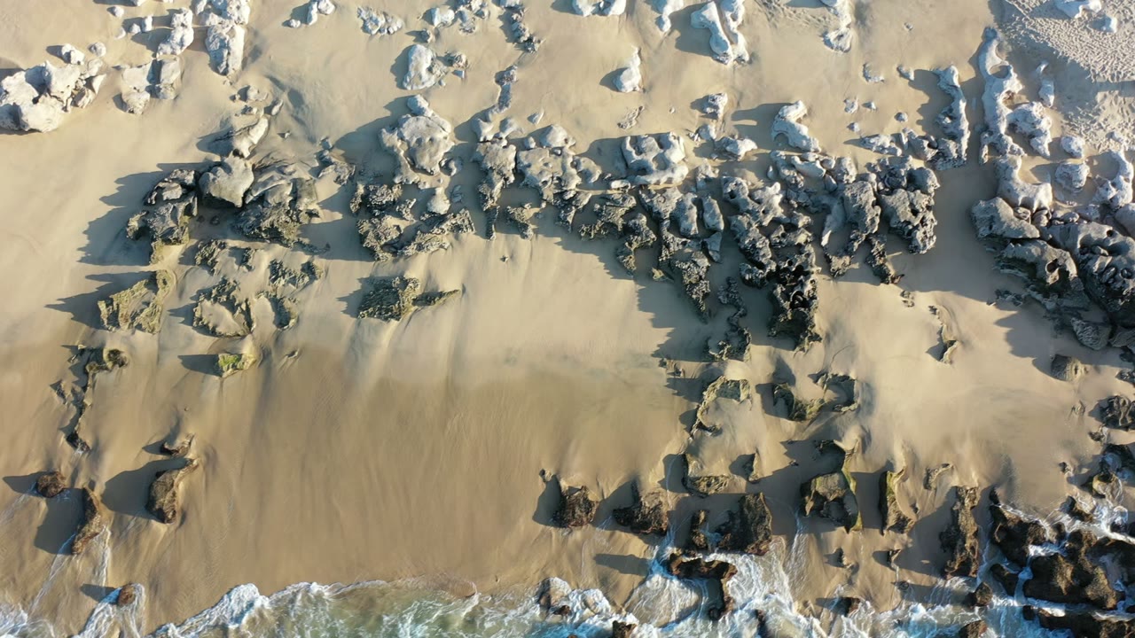 Top-down view of a Los Cabos sandy beach with dark rock bands, scattered pale stones, small surf at the bottom edge, and no visible people.