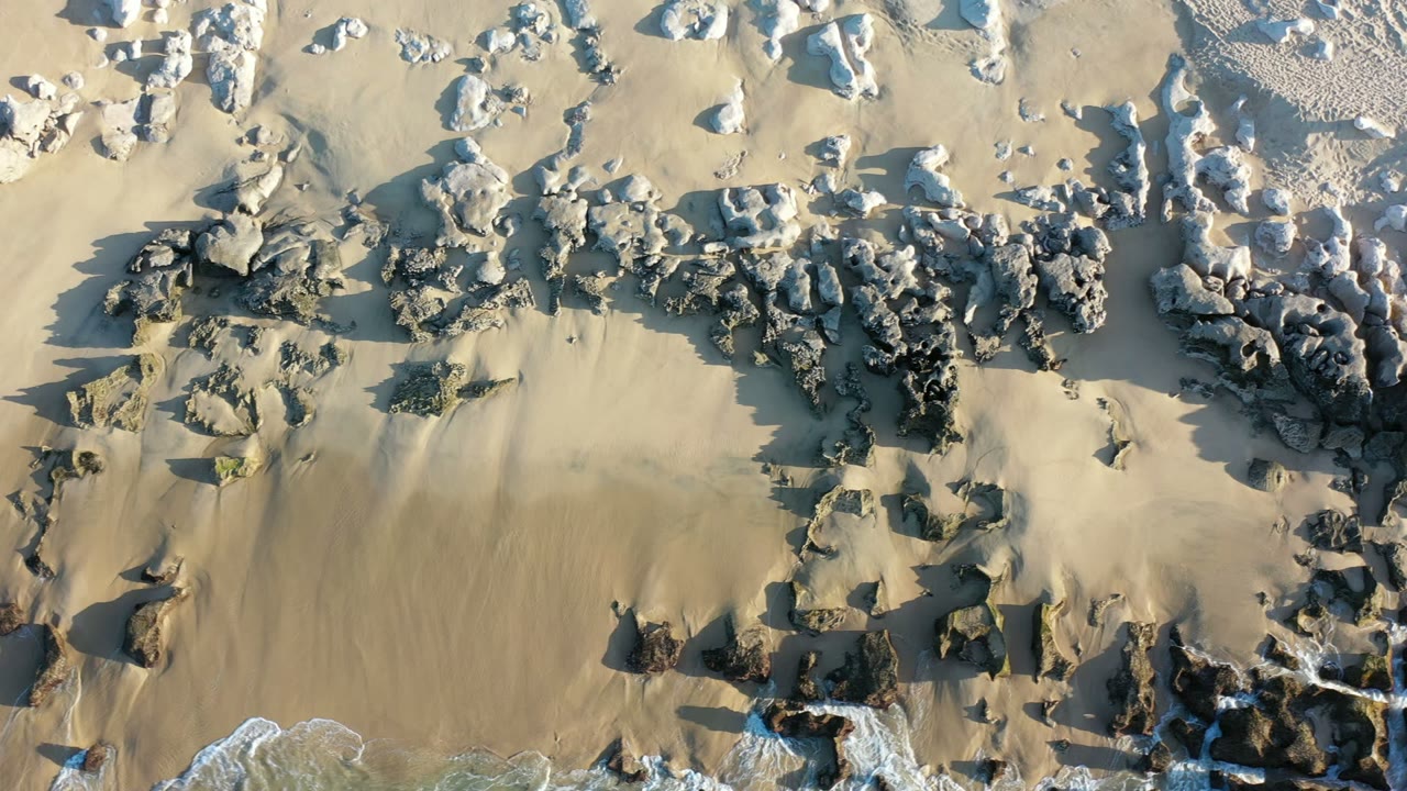 Aerial Los Cabos shoreline frame with clustered dark rocks across the center, pale sand around them, a thin white wave line below, and no visible people.