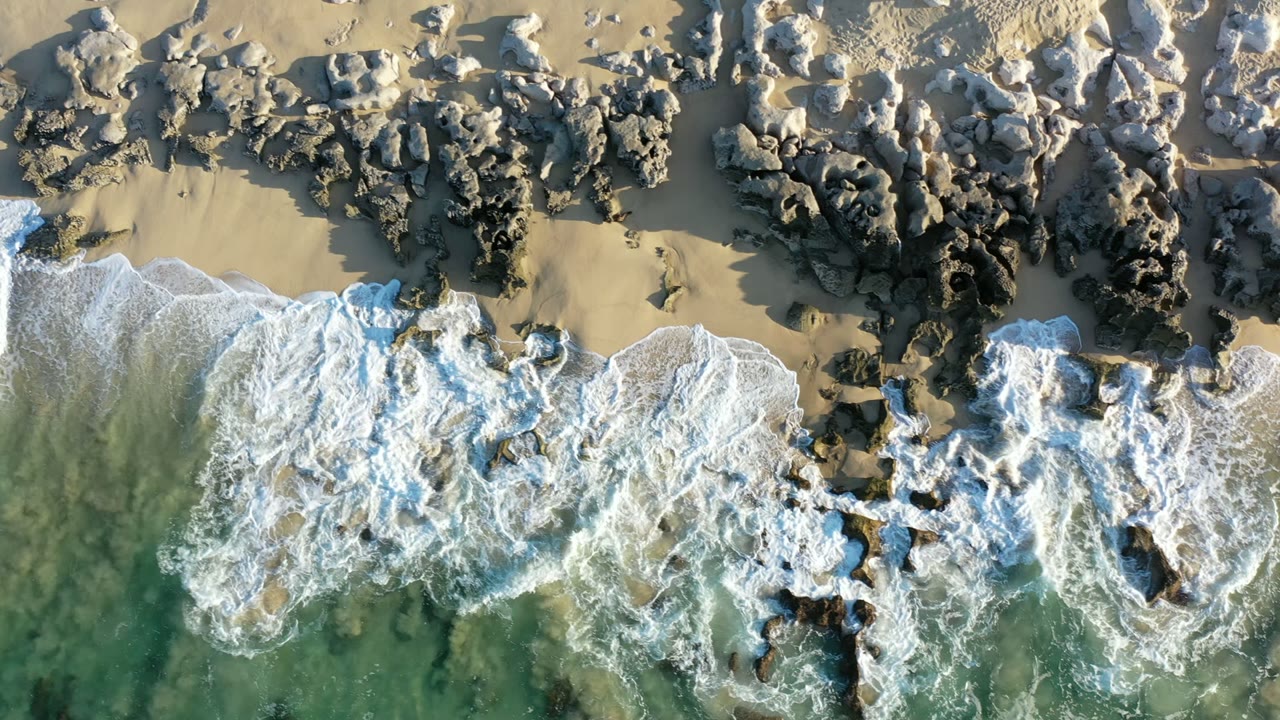 Aerial frame over Los Cabos, Mexico with a larger band of white water breaking across clustered rocks and tan sand, with no visible people.