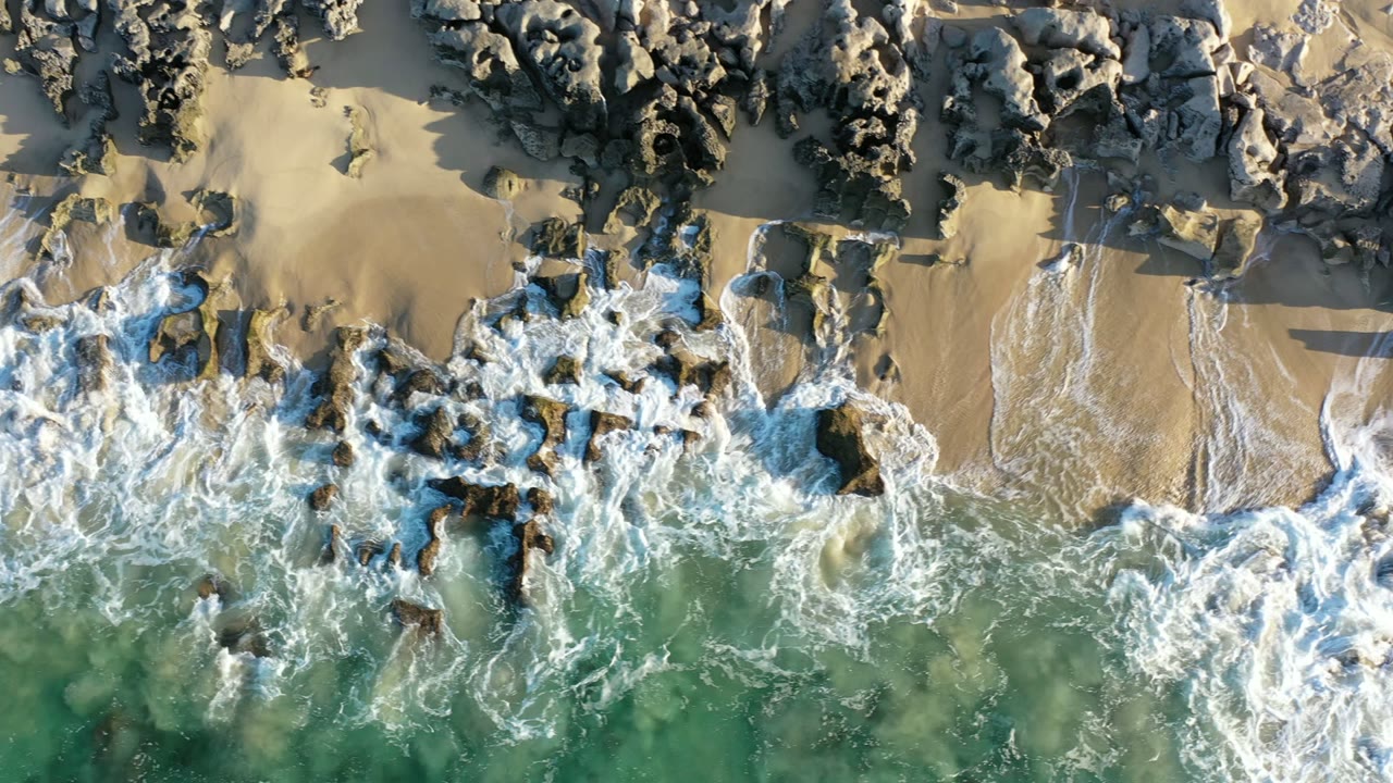 Overhead view in Los Cabos, Mexico of white surf breaking around dark rocks on tan sand, with green-blue water below and no visible people.
