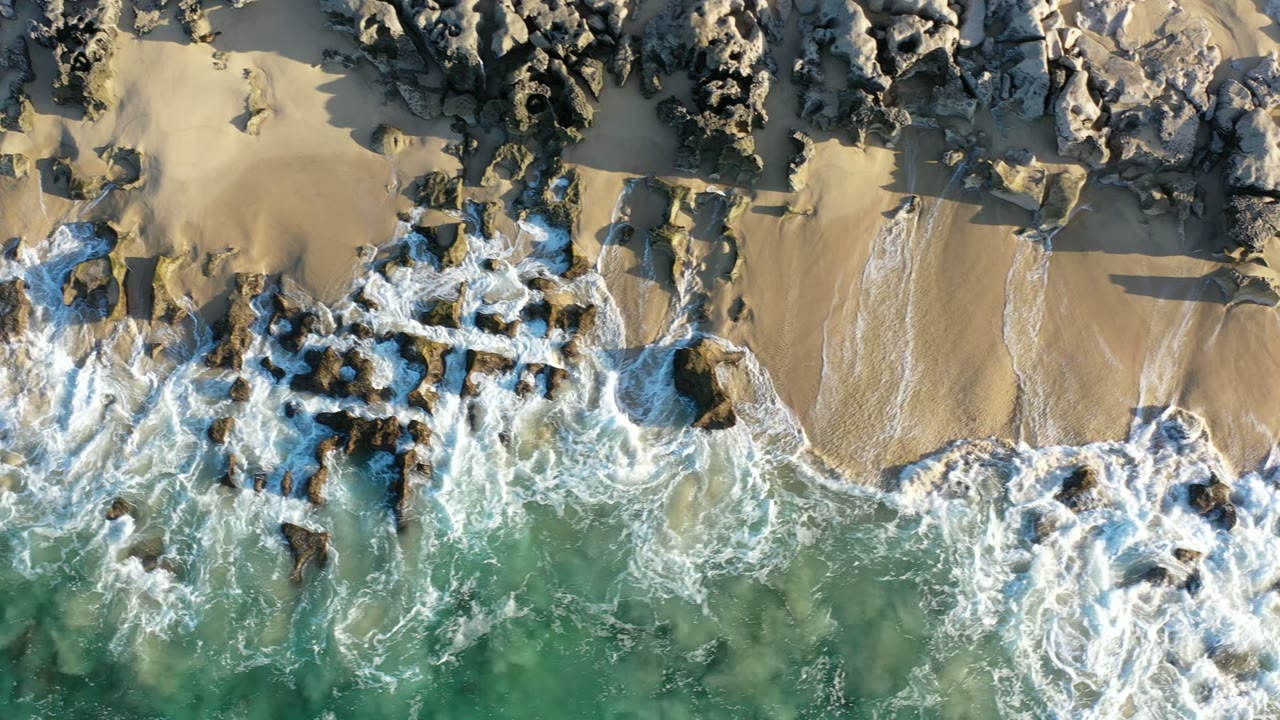 Overhead Los Cabos shoreline frame with foam spreading across clustered rocks, wet sand channels, green-blue water, and no visible people.