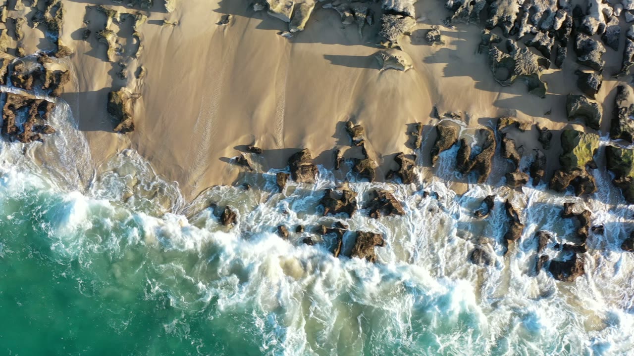 Overhead Los Cabos shoreline frame with a broader band of white foam moving across scattered dark rocks below smooth tan beach, with no visible people.