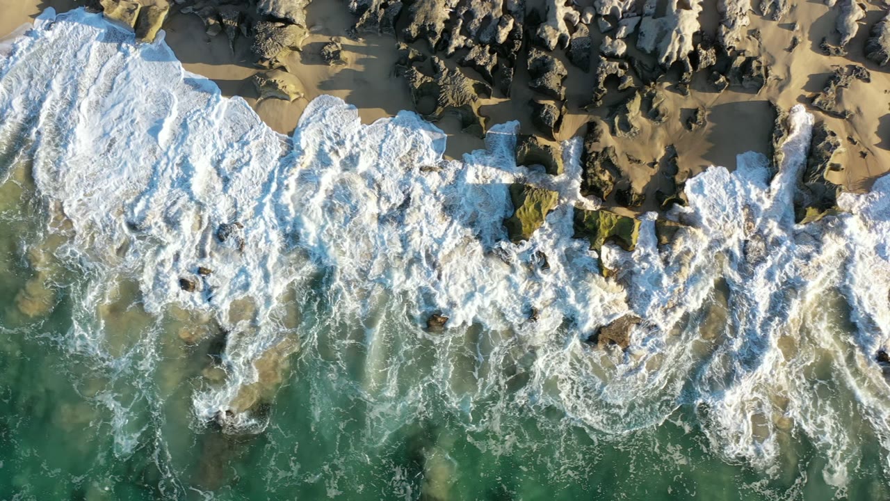 Overhead view in Los Cabos, Mexico of white surf spreading across dark shoreline rocks and tan sand, with green water below and no visible people.