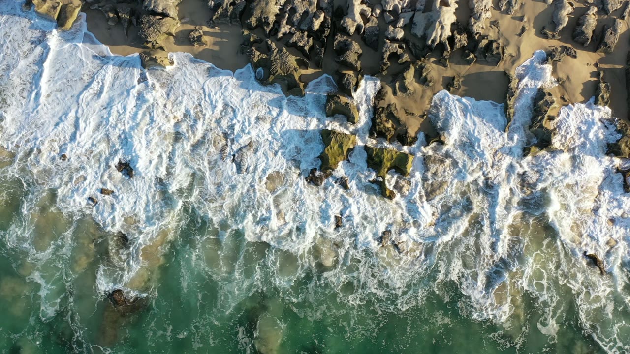 A second overhead moment in Los Cabos, Mexico shows foamy waves wrapping around scattered rocks, with a strip of dry sand at the top and no visible people.