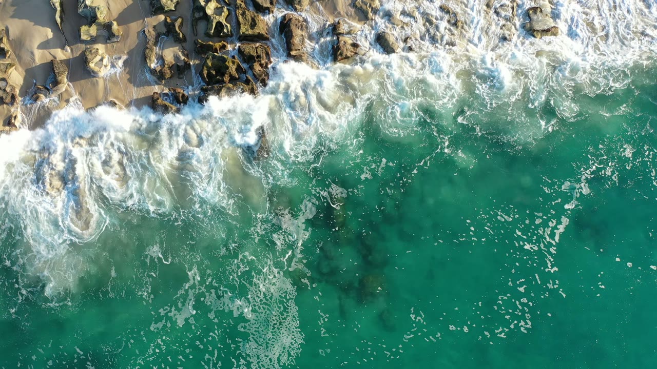 Aerial frame in Los Cabos, Mexico, where a larger band of white foam breaks across tan sand and scattered rocks above green-blue water with no visible people.