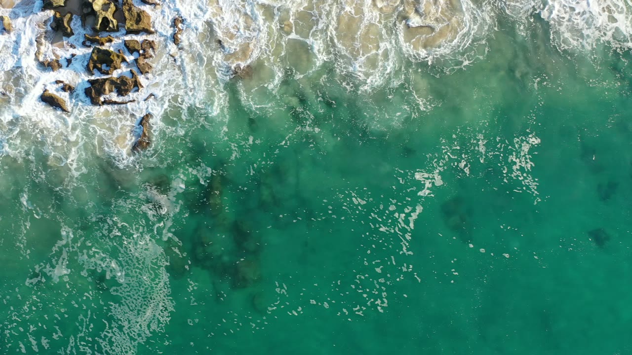 Overhead view in Los Cabos, Mexico, showing turquoise water below white surf wrapping around brown rocks near the upper edge, with no visible people.