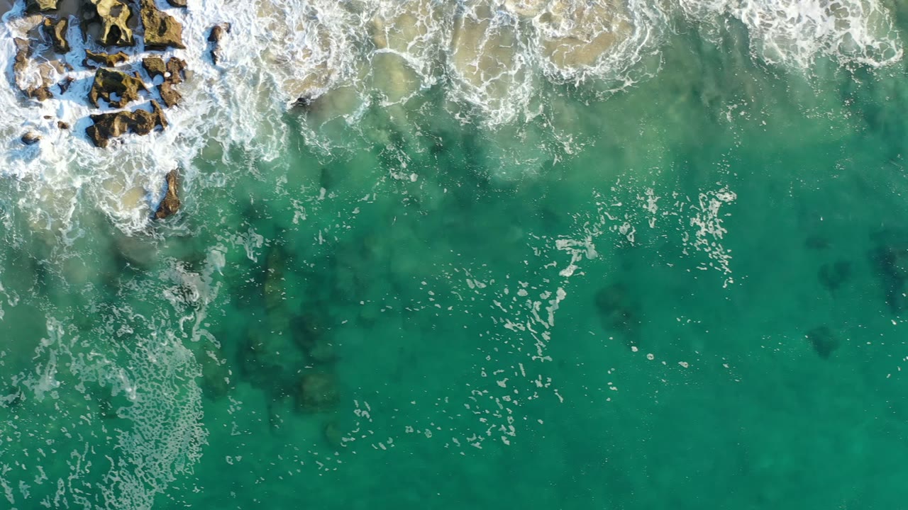 Aerial frame from Los Cabos, Mexico, with white wave foam spreading downward from brown rocks into green-blue shallow water, with no visible people.