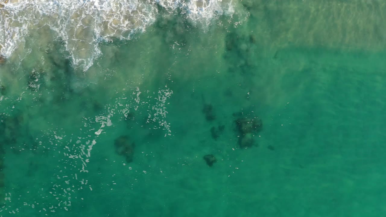 Aerial Los Cabos shoreline frame with a foamy wave line across the upper edge, green-blue water below, scattered dark underwater shapes, and no visible people.