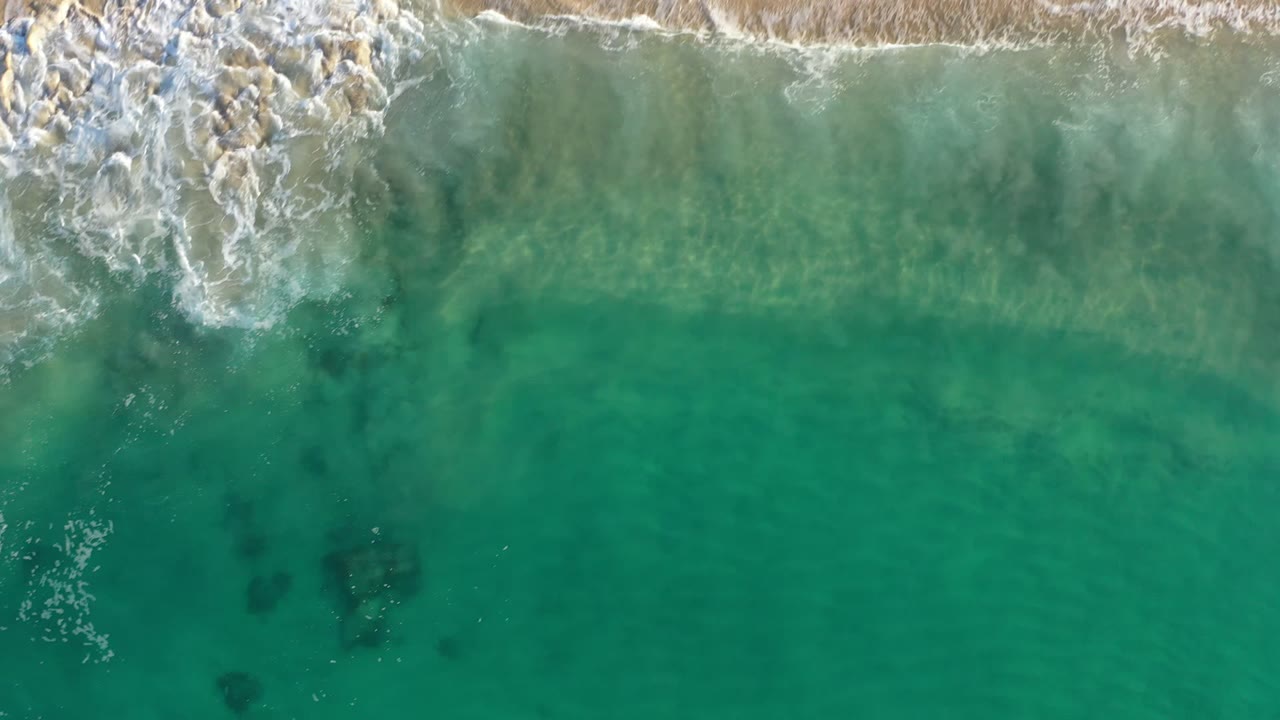 Overhead Los Cabos beach frame with a stronger band of white shore break across the top and clear green-blue water below, with no visible people.
