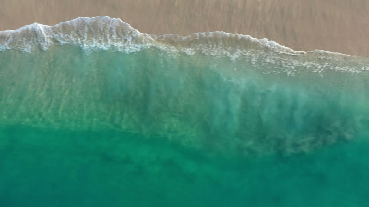 Overhead view in Los Cabos, Mexico, showing green sea below a strip of pale sand and foamy surf, with no visible people.