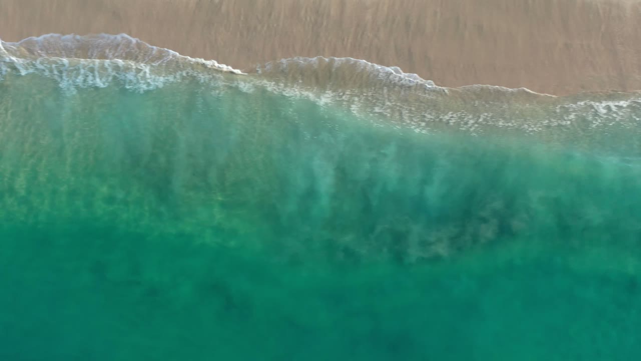 Aerial Los Cabos water view with turquoise-green sea dominating the frame and a thin white wave line along the sandy beach, with no visible people.