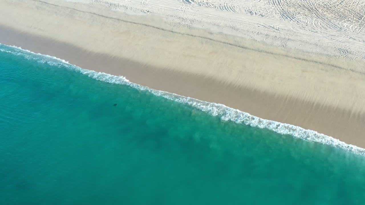 Aerial Los Cabos shoreline frame with the wave edge cutting diagonally across turquoise water and smooth pale sand marked by faint tracks near the top.