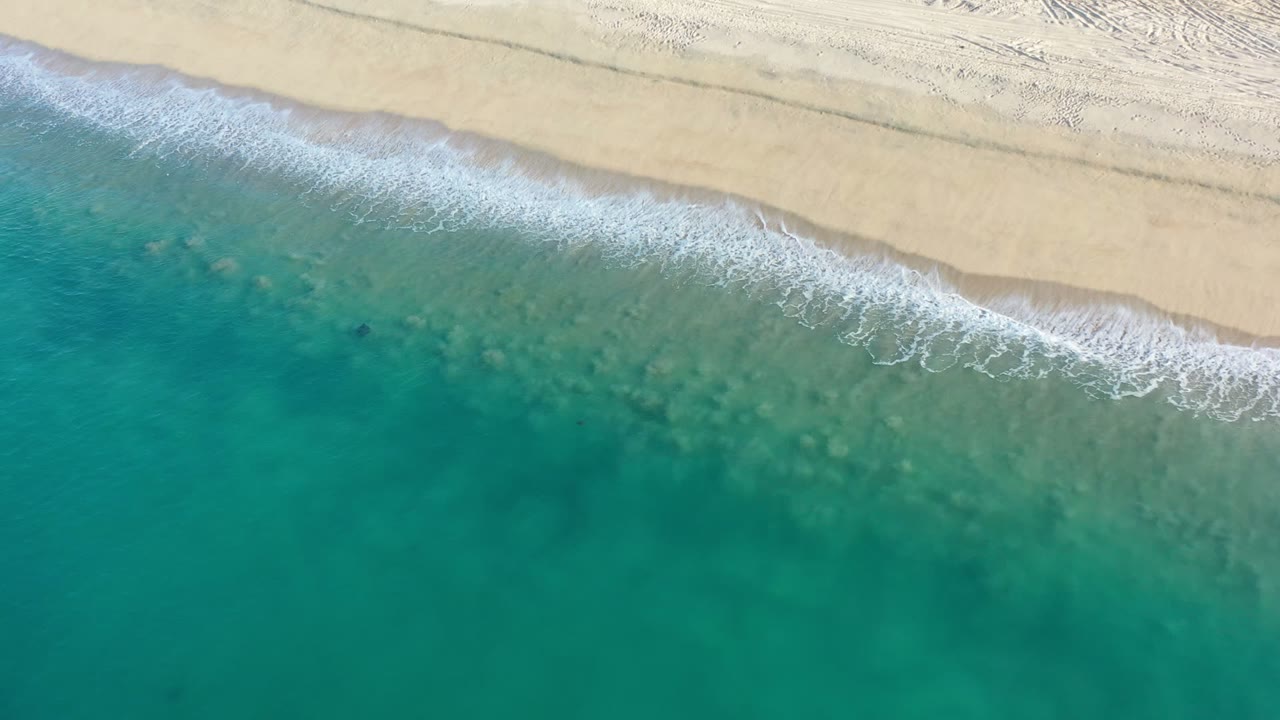 Aerial frame of white shore break tracing the edge of an empty Los Cabos beach, with green water below and pale sand above, no visible people.