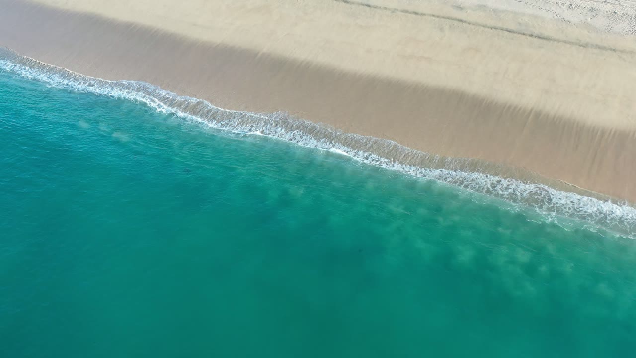 Overhead view of turquoise ocean reaching pale sand in Los Cabos, Mexico, with a narrow white foam edge and no visible people.