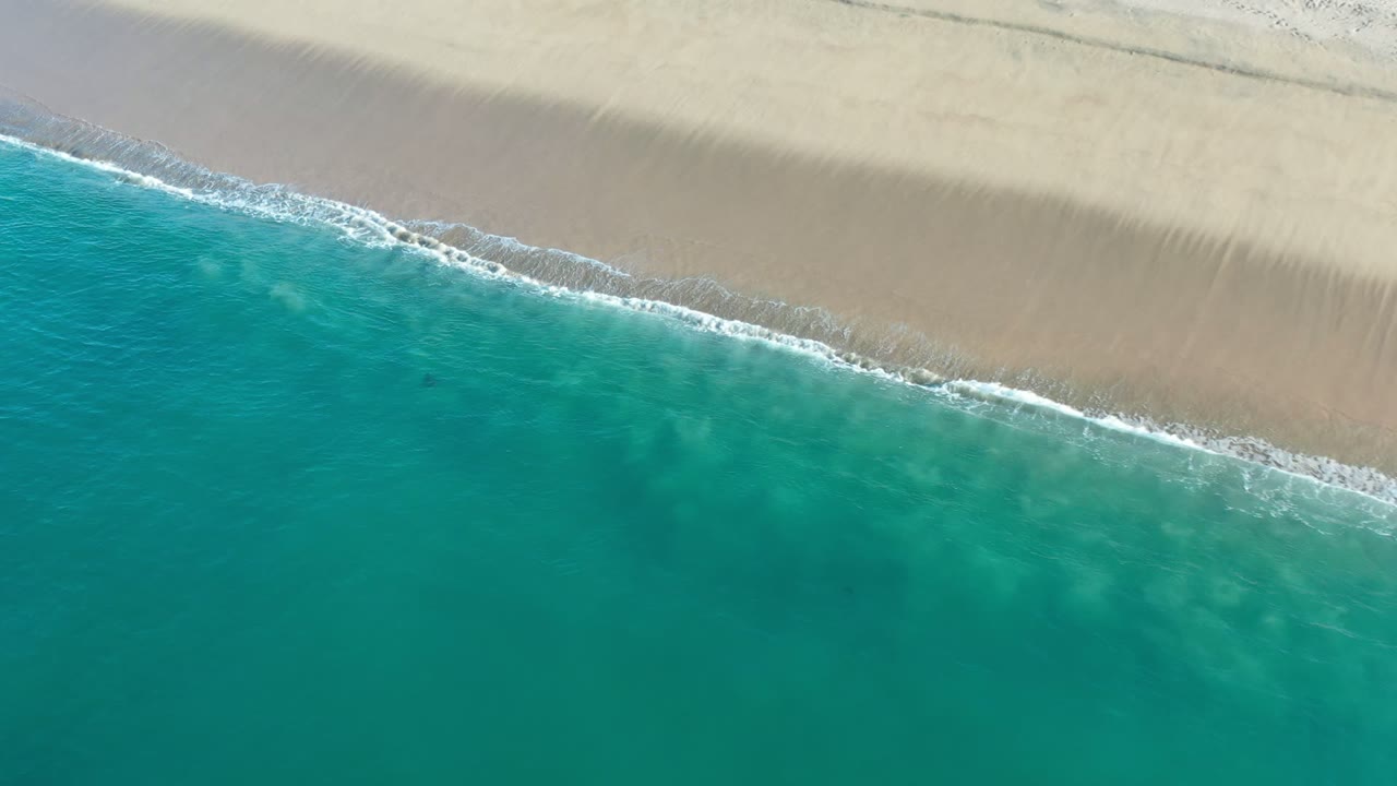 Aerial frame of clear teal water beside pale Los Cabos sand, with the surf line slightly farther across the frame and no visible people.