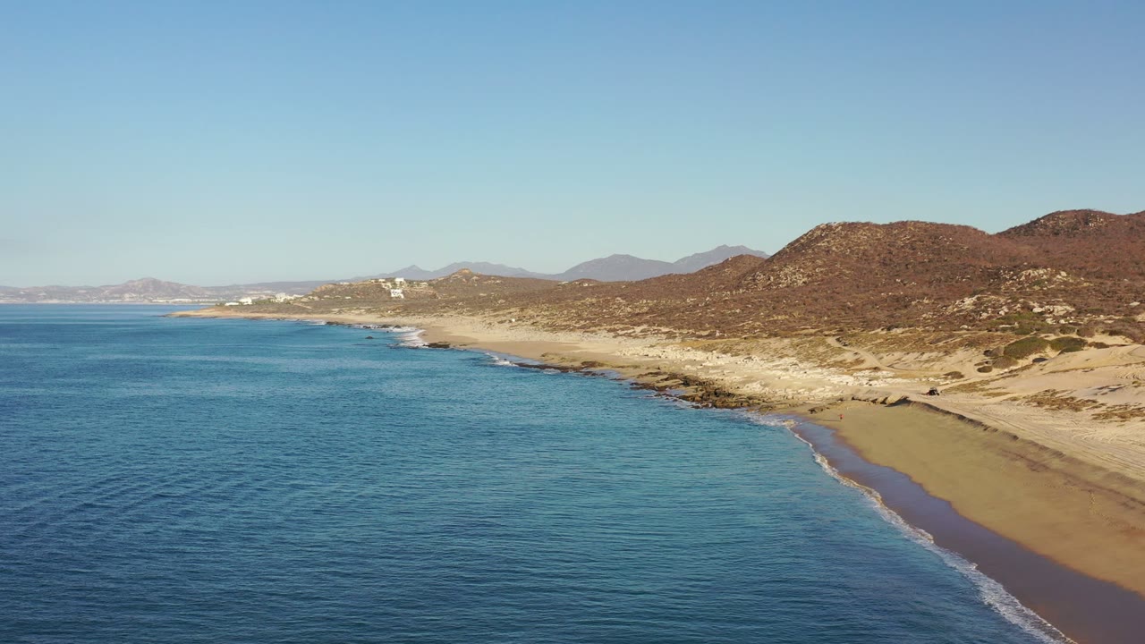 Aerial coastline with blue ocean, sandy beach, dry hills, and distant mountains.