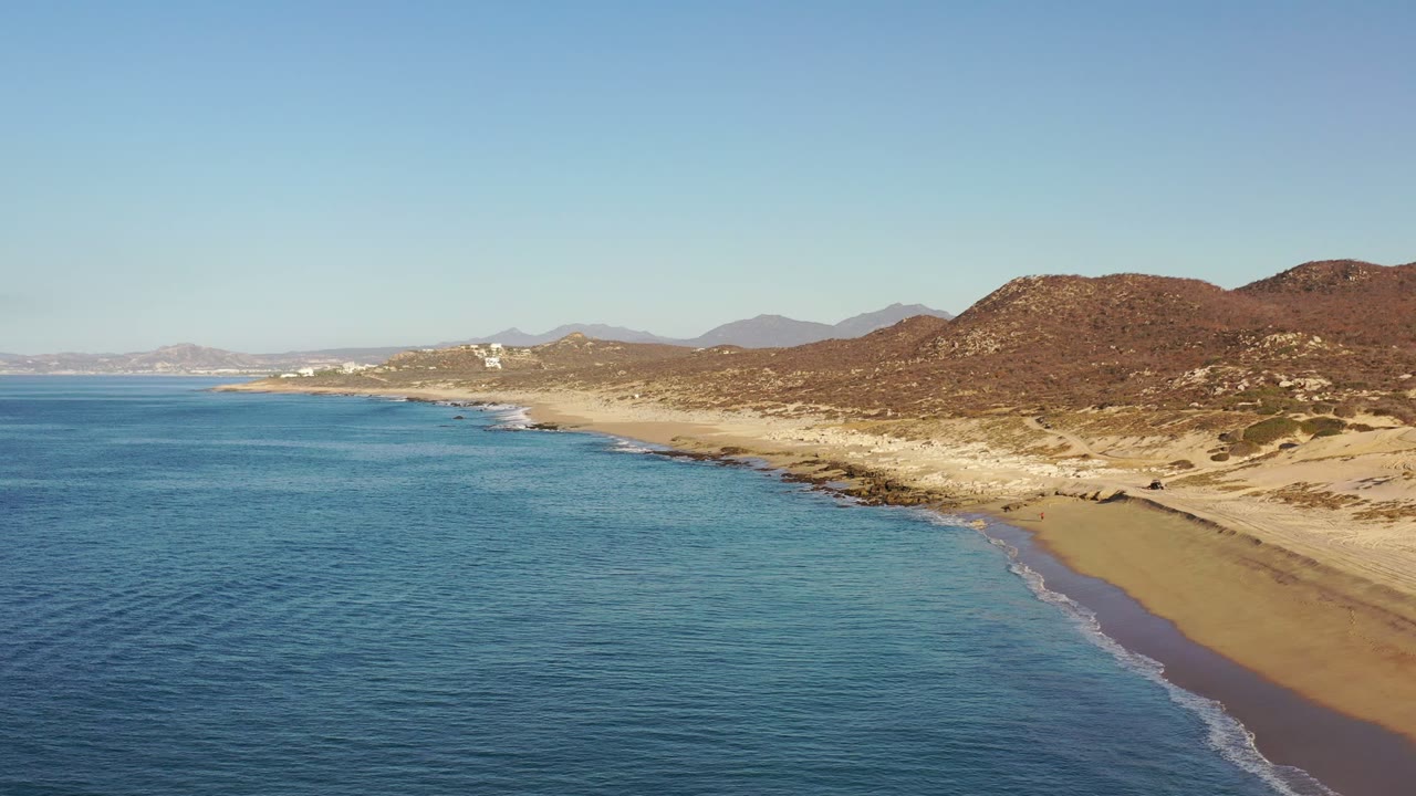Aerial Los Cabos beach view one second in, with a thin white surf edge tracing the sand, scrubby brown hills inland, and no visible people.