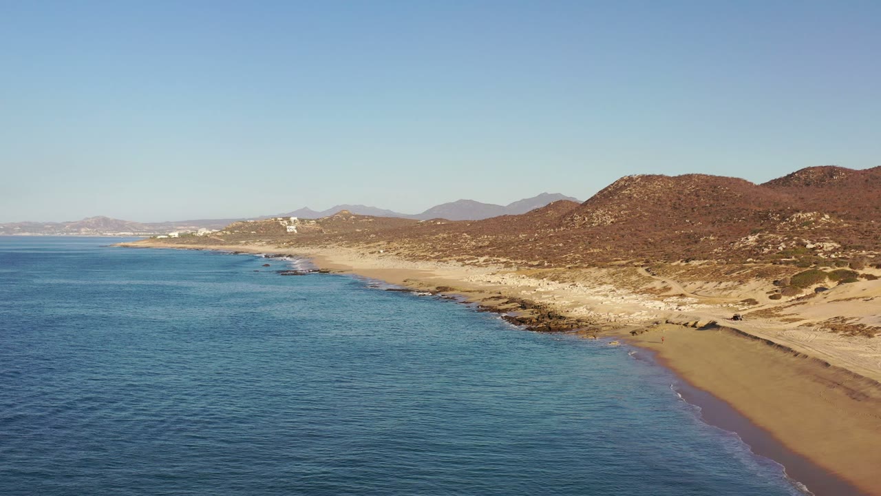 Aerial view of blue ocean meeting a sandy Los Cabos beach with low brown hills beyond.