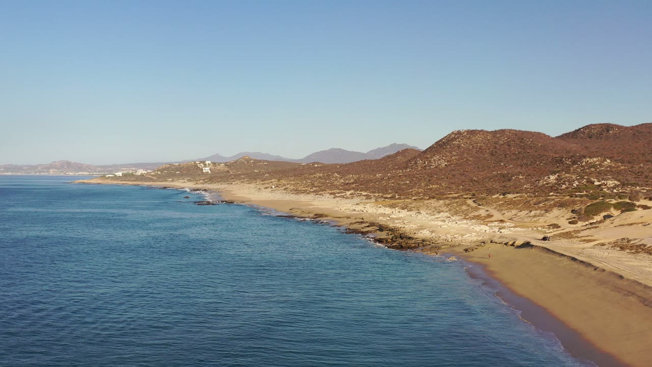 Los Cabos coastline frame with the ocean filling the left half, a narrow surf line along the sand, rocky shoreline ahead, brown hills, and no visible people.