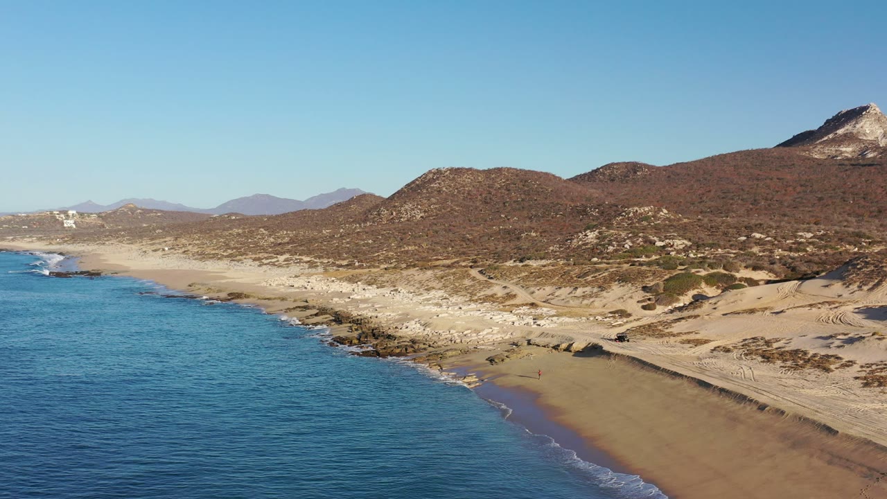 Aerial view in Los Cabos, Mexico with blue ocean at left, pale sand shoreline, rocky coast, dry hills, and a pointed mountain in the distance, with no visible people.