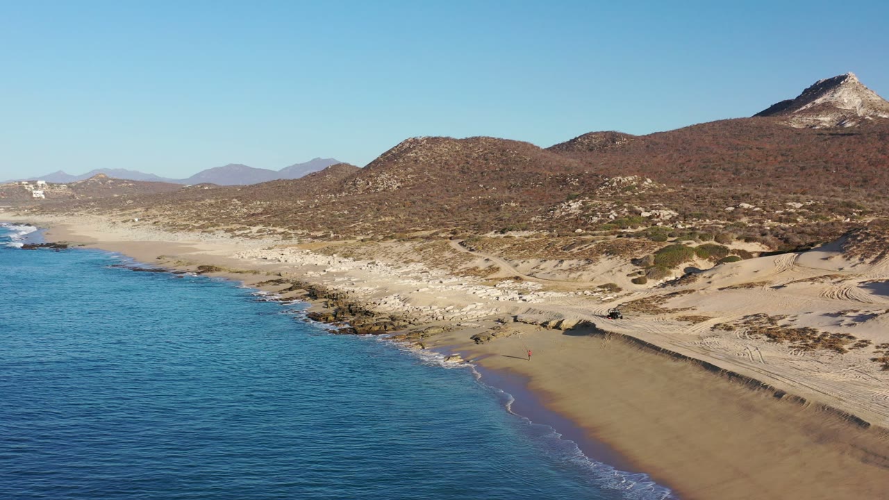 Aerial frame over Los Cabos, Mexico showing rippled blue water, a curved rocky shore, pale beach sand, scrub-covered hills, and distant mountains, with no visible people.
