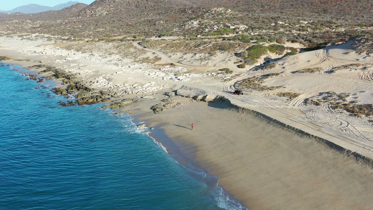 Aerial view over a Los Cabos, Mexico beach with turquoise water at left, broad sand at center, dunes and rocky scrubland beyond, and no clearly identifiable people.