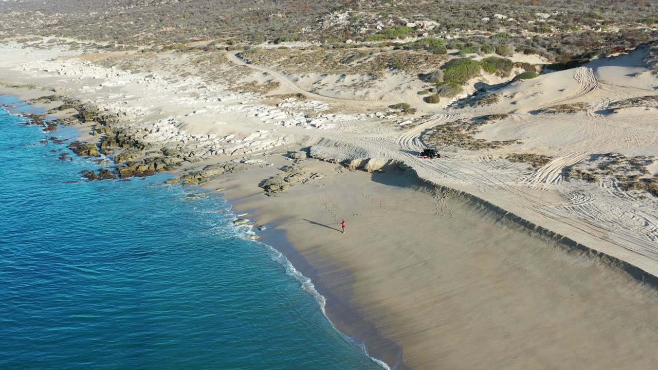Aerial frame of a Los Cabos beach where shallow blue water curls onto wet sand, with pale dunes, rocky patches, vehicle tracks, and a tiny distant red figure on the beach.