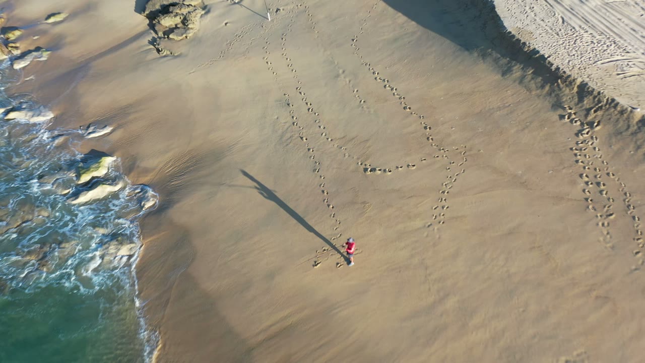 Overhead view of a Los Cabos beach showing foamy surf at left, tan sand, scattered rocks, footprints, and a tiny red-clad person casting a long shadow.