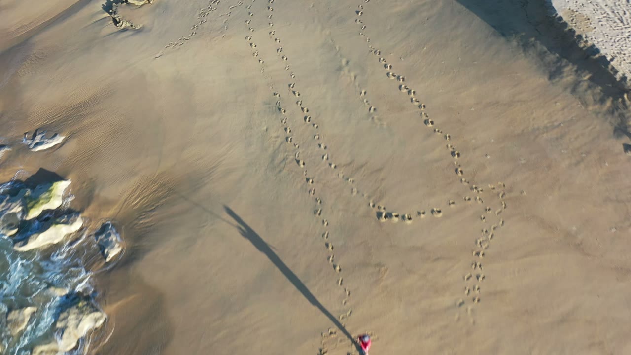 Top-down Los Cabos beach frame with broad tan sand, footstep trails, pale rocks, a long diagonal shadow, and a tiny red figure near the bottom edge.