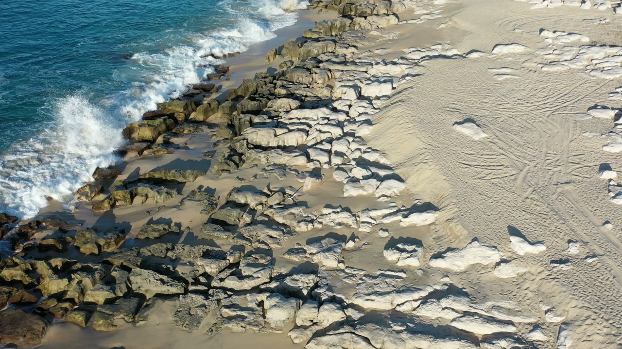 A wave pushes white foam farther into the rock line on the Los Cabos beach, with textured sand and scattered stones visible to the right and no visible people.