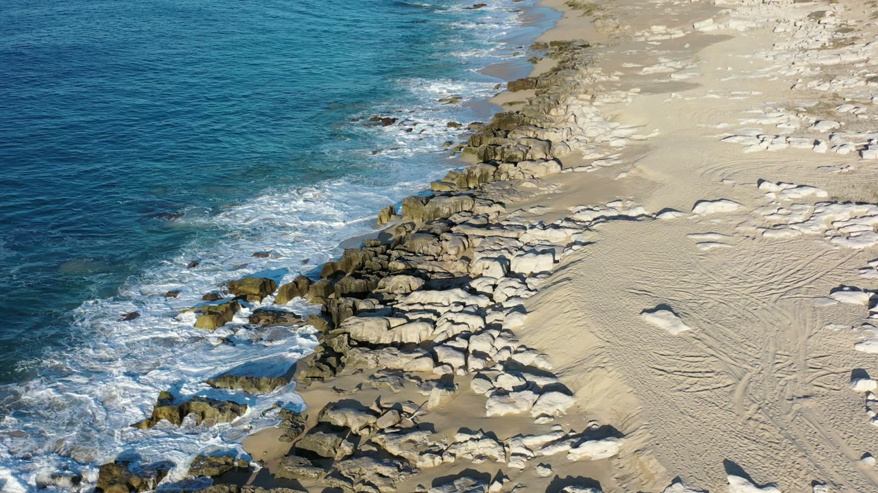 Aerial view of waves breaking on rocks beside a sandy beach.