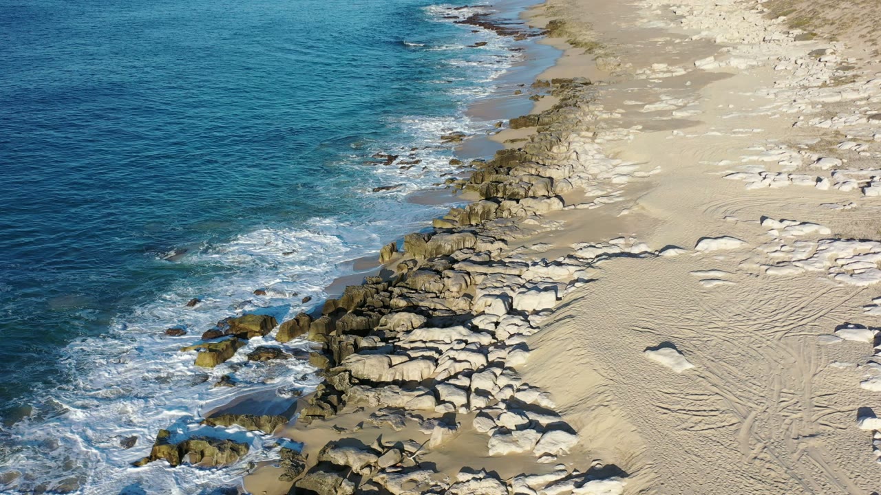 Aerial coastal frame in Los Cabos, Mexico showing foamy waves wrapped around a cluster of rocks with rippled sand to the right and no visible people.