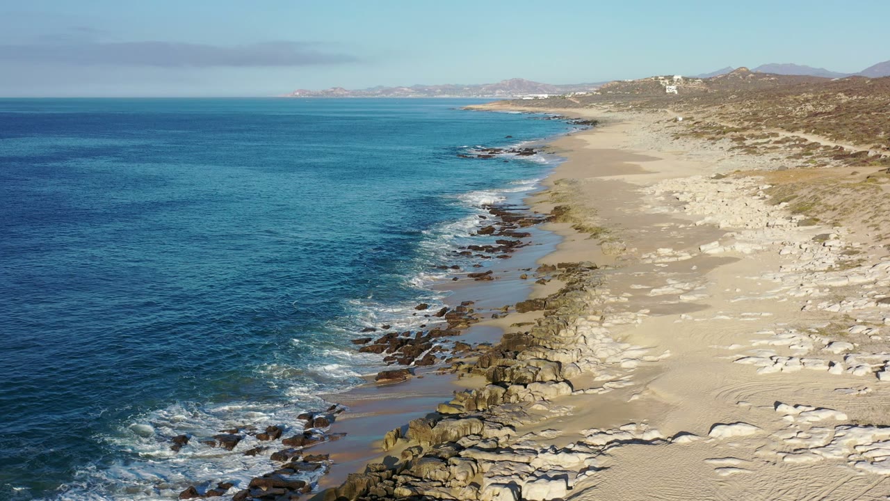 Opening frame of the Los Cabos, Mexico coastline with blue ocean on the left, white surf touching rocks and sand, scrubby hills to the right, and no visible people.