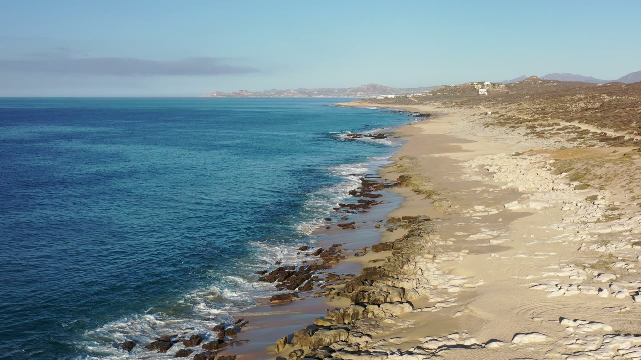 Second frame over the Los Cabos, Mexico beach, where small waves break against darker shoreline rocks below a pale sandy stretch and no visible people appear.