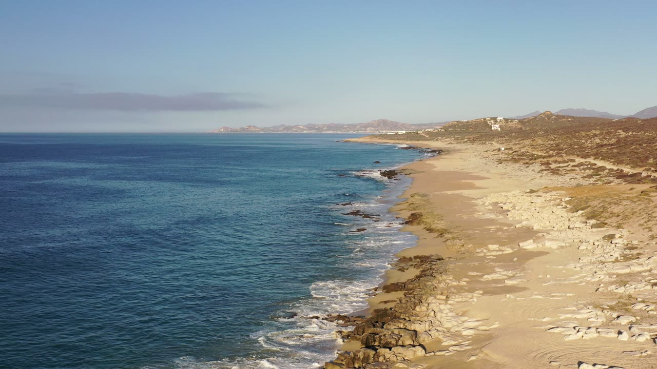 Aerial coastline with ocean, sandy beach, rocks, and distant hills.