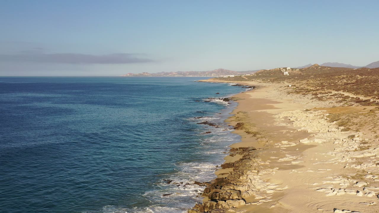 Elevated Los Cabos, Mexico shoreline view with the ocean occupying the left side, wave foam along the beach edge, dry scrubland to the right, and no visible people.