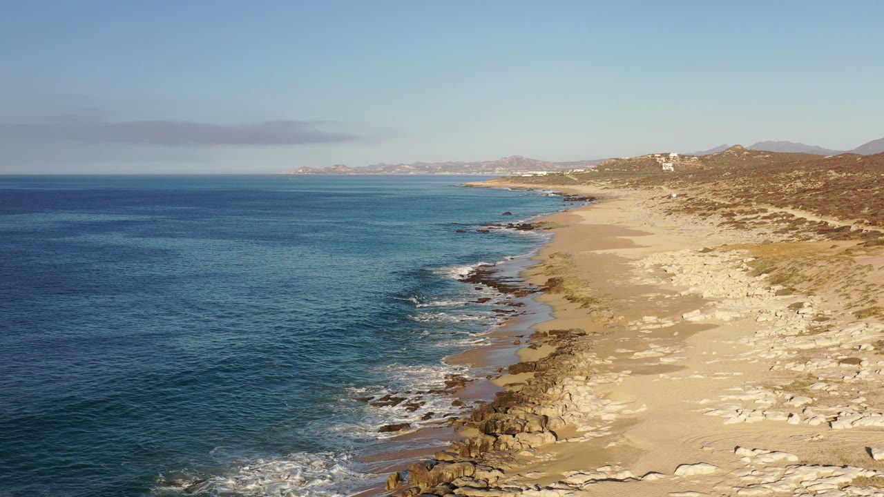 Elevated view of the Los Cabos, Mexico coast with blue water on the left, pale sand and rocks on the right, distant hills, and no visible people.