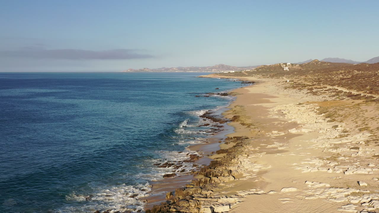 Aerial coastal view in Los Cabos, Mexico showing small waves breaking along dark rocks, dry sand with faint tire marks, scrubland, and no visible people.