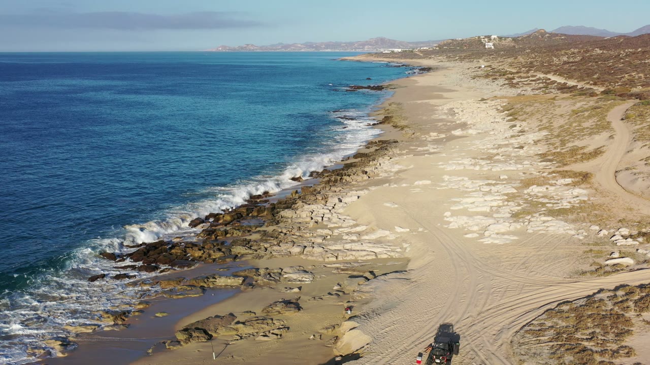 Elevated Los Cabos shoreline view with white surf breaking over dark rocks, turquoise shallows, wide sand, sparse scrub, and no visible people.