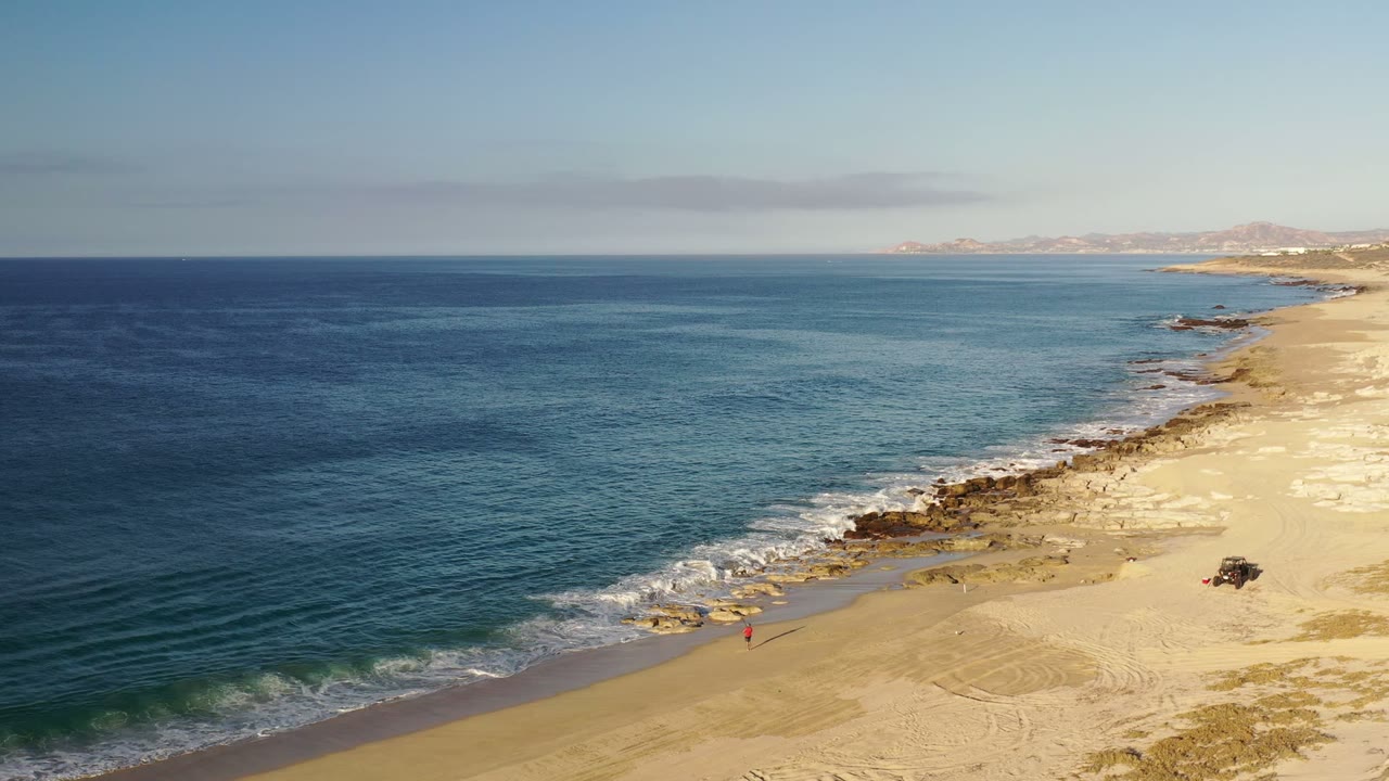 Los Cabos, Mexico beach from above, with small white waves crossing the sand, darker rocks near the waterline, distant hills on the horizon, and no visible people.