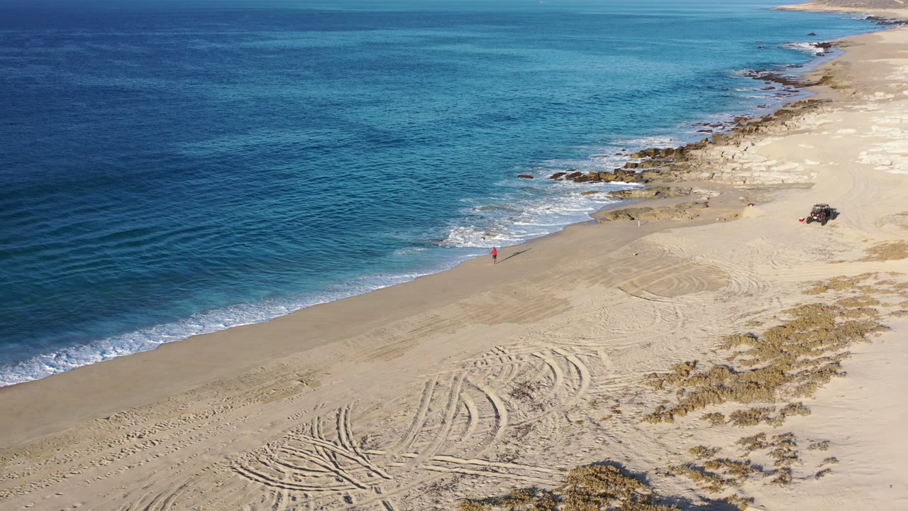 Aerial view of a Los Cabos beach at 0:00, with deep blue water on the left, pale sand below, curved tire tracks in the foreground, and a tiny red figure near the waterline.
